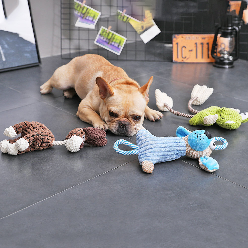 French bulldog lying on a dark tile floor surrounded by several squeaky plush dog toys, including a blue puppy, a brown hedgehog, and a green frog