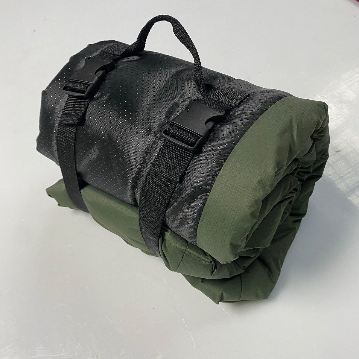 Real-world photo of the military-green roll-up dog bed rolled up with its black perforated cover, straps, and carry handle on a metallic surface.