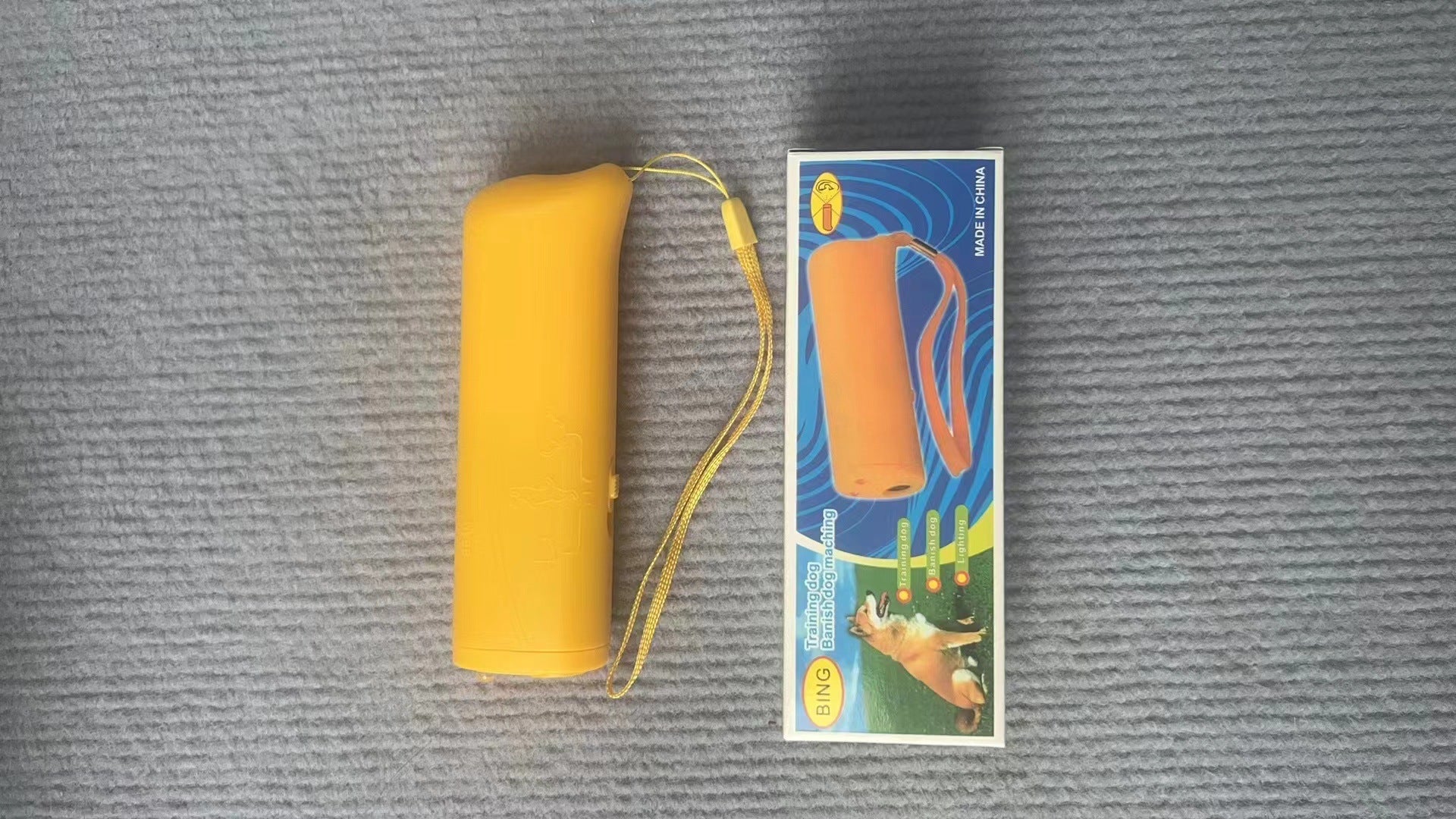 Real-world photo of the yellow ultrasonic dog trainer with its wrist strap lying on a gray surface next to its printed retail box.