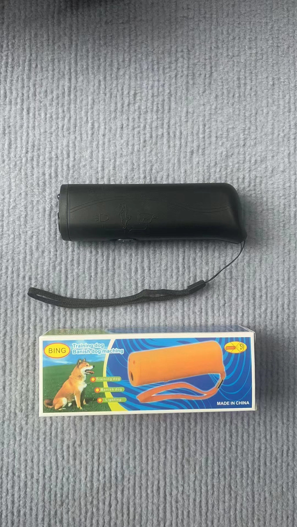 Real-world photo of the black ultrasonic dog trainer and its wrist strap on a gray surface beside its printed retail box.