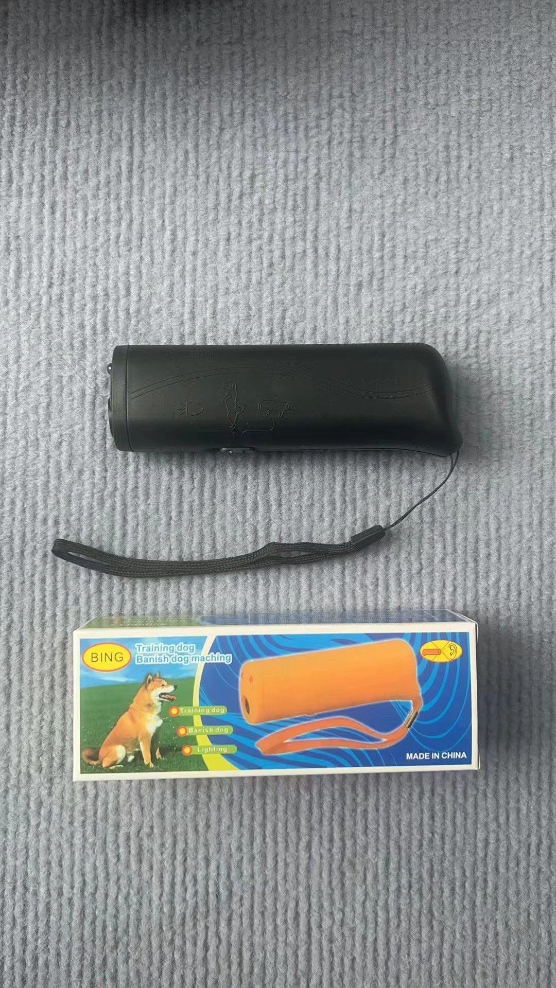 Real-world photo of the black ultrasonic dog trainer and its wrist strap on a gray surface beside its printed retail box.