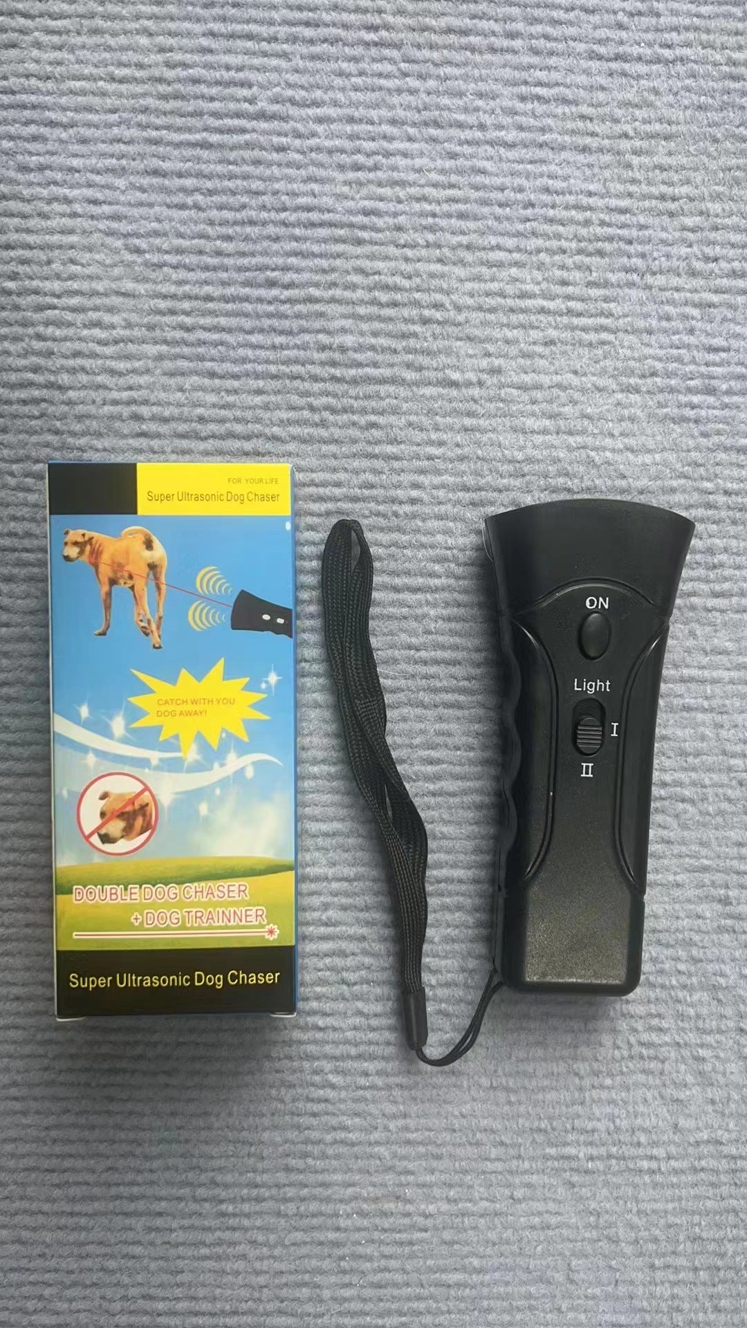 Real-world photo of the black ultrasonic dog trainer with its retail box and detached wrist strap arranged on a gray surface.